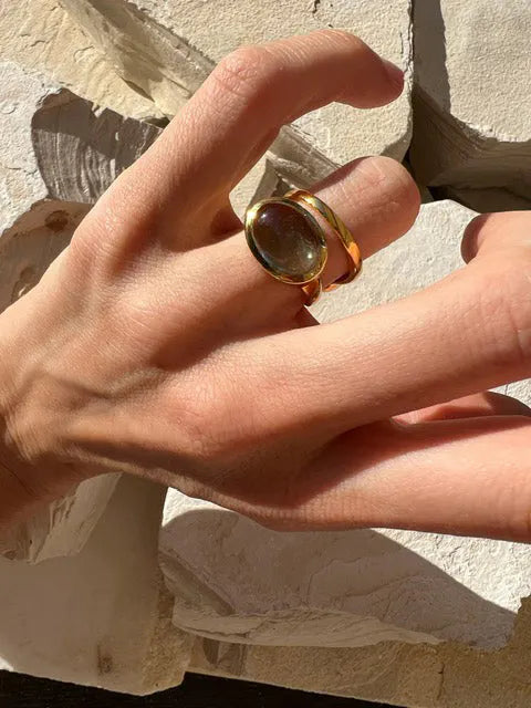 Close-up of a hand in sunlight, wearing a large, rounded, translucent Sian Gold Plated Ring by Shyla with a 22 carat gold band. The background consists of light-colored, rough-textured stones or tiles. Shadows emphasize the texture and shape of both the hand and the ring.