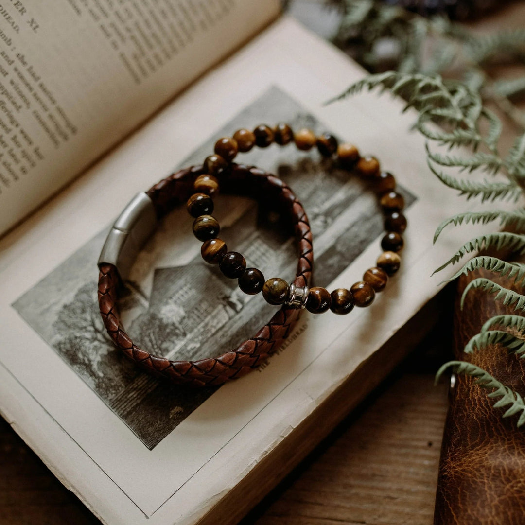 Two bracelets sit atop an open book with an illustration on one page. One is a Steel and Barnett "Preston - Caramel" braided leather bracelet crafted from genuine brown leather with a metal clasp, and the other consists of dark-colored round beads. Fern leaves partially frame the scene, adding to the rustic and warm setting.