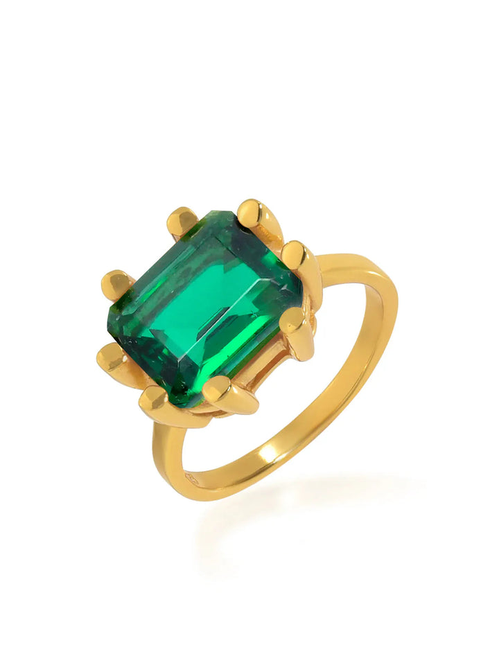 A chunky ring featuring a large rectangular-cut emerald green gemstone set in 22 carat gold. The square stone is held in place by four gold prongs, and the ring has a simple, elegant band. Displayed against a plain white background, this is the Square Claw Gold Plated Ring by Shyla.