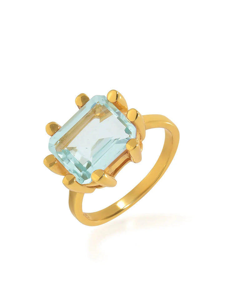 A chunky ring crafted from 22 carat gold, featuring a large, rectangular-cut light blue gemstone set in a prong setting. The smooth, shiny band perfectly complements the gemstone's prominent position, catching the light and highlighting its breathtaking facets. This is the Square Claw Gold Plated Ring from Shyla.