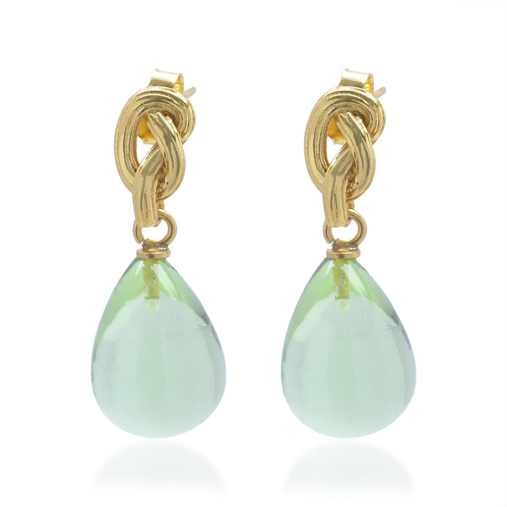 The Shyla Synthea Earrings in various colours feature an intricate gold knot top with a large, translucent light green teardrop gemstone gracefully dangling below. Hypoallergenic for sensitive ears.