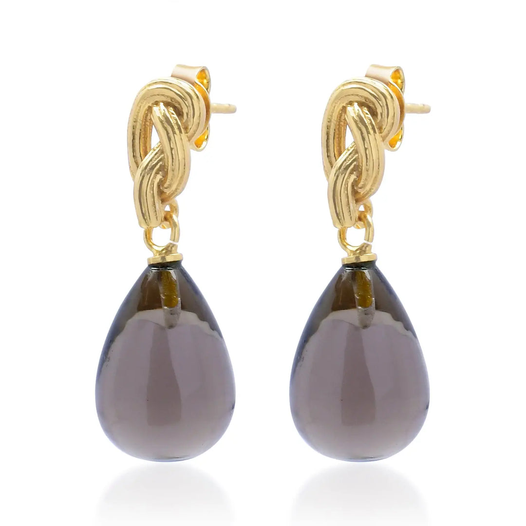Elegant and timeless, the Synthea Earrings in dark gray from Shyla feature large, smooth, teardrop-shaped gemstones gracefully suspended below intricate 22-carat gold knot studs. Displayed against a white background, these hypoallergenic earrings offer both sophistication and comfort for all-day wear.