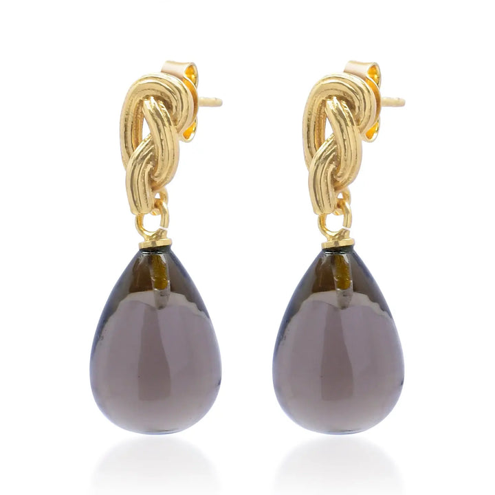 Elegant and timeless, the Synthea Earrings in dark gray from Shyla feature large, smooth, teardrop-shaped gemstones gracefully suspended below intricate 22-carat gold knot studs. Displayed against a white background, these hypoallergenic earrings offer both sophistication and comfort for all-day wear.