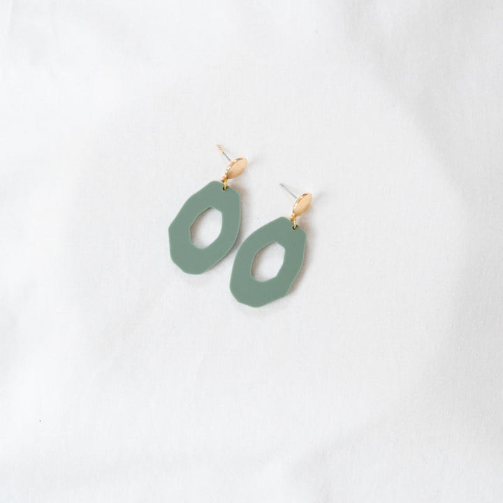 A pair of stylish, handmade earrings lies on a white fabric background. Each earring features an irregular oval-shaped green resin piece made from recycled acrylic with a geometric cutout in the center, connected to a small, gold-colored stud. The design is modern and minimalist, perfect for those who love lightweight earrings. These are the Sage Green Irregular Oval Acrylic Gold Woods Earrings by Studio Nok Nok.