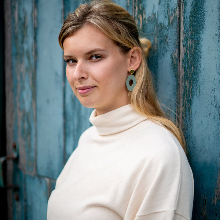 A person with long blonde hair stands in front of a weathered, blue wooden door. They are wearing a cream-colored turtleneck sweater and a pair of large, lightweight Sage Green Irregular Oval Acrylic Gold Woods Earrings by Studio Nok Nok. They have a slight smile and are looking directly at the camera.