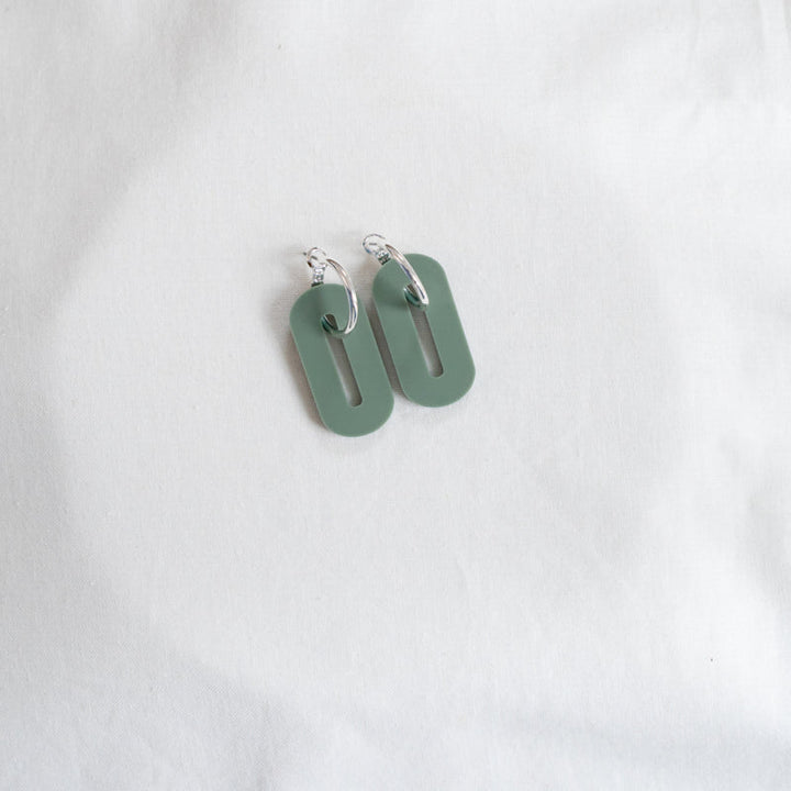 A pair of Studio Nok Nok Sage Green Oval Misty Acrylic Silver Hoop Earrings on a white fabric background. Each earring features a silver hoop and a vertically oriented, rectangular, green pendant made from recycled acrylic with rounded edges and an oval cutout in the center.