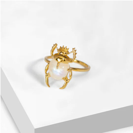 A Marie June Jewellery Sacred Scarab Gemstone Ring designed as a sacred scarab beetle, featuring detailed legs and antennae. The beetle's body is formed by a large, polished white pearl. The ring is displayed on a white geometric platform, with a minimalistic background.