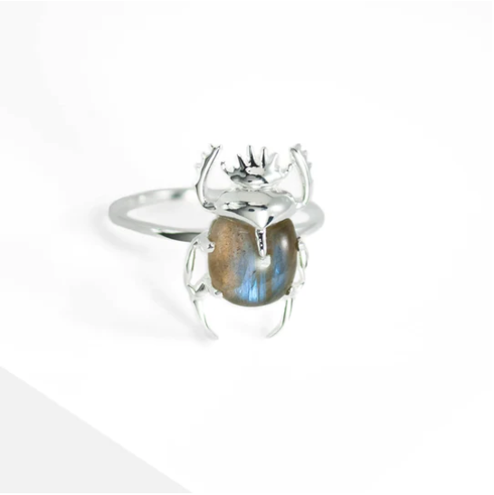 A silver Sacred Scarab Gemstone Ring from Marie June Jewellery designed to resemble a beetle, holding a polished blue gemstone in its body. The beetle's legs and antennae are intricately detailed. The background is white.