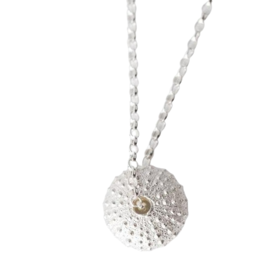 The Jennifer Kinnear Sea Urchin Necklace Small, available in silver or gold, features a textured sea urchin-inspired pendant with a central circular accent on a simple chain, displayed against a white background.