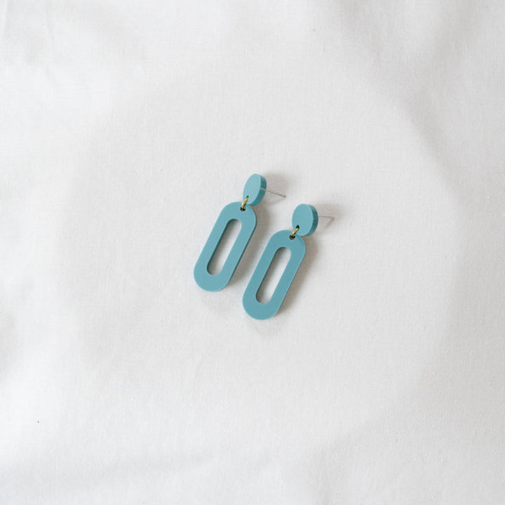 A pair of light blue, modern, geometric Timeless 04 Oval Acrylic Earrings from Studio Nok Nok placed on a white fabric background. These perfect earrings feature an oval-shaped stud connected to a larger, vertically elongated open rectangle shape, and they're crafted from recycled materials.