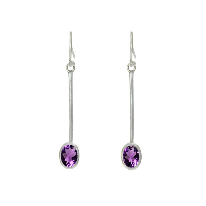 The Sequola Earrings by Gallardo & Blaine Designs feature long sterling silver hooks with dangling rods, each showcasing a single faceted oval gemstone in purple. The plain white backdrop enhances their elegance and timeless charm.