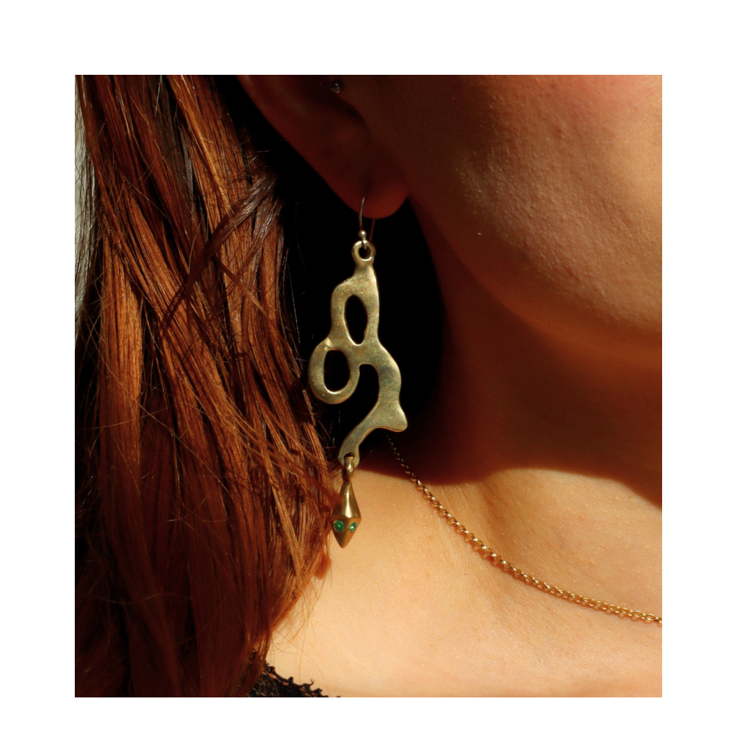 A close-up of a person with long brown hair wearing Madeleine Sculptural Jewellery’s Serpent Earrings—large, abstract-shaped gold pieces with organic lines—against a white background, highlighting the beauty of handmade design.
