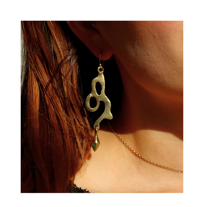 A close-up of a person with long brown hair wearing Madeleine Sculptural Jewellery’s Serpent Earrings—large, abstract-shaped gold pieces with organic lines—against a white background, highlighting the beauty of handmade design.
