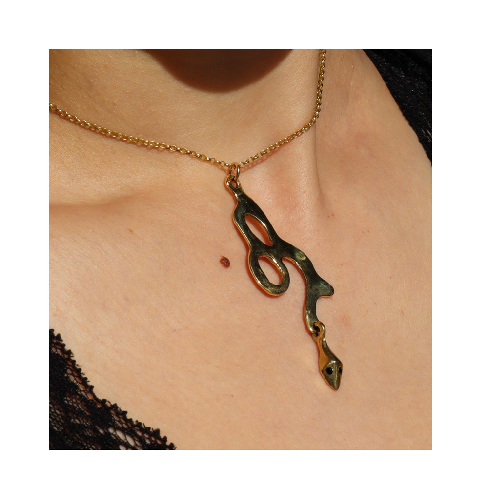 The Serpent Pendant by Madeleine Sculptural Jewellery is a gold chain necklace featuring an abstract, irregular pendant made from recycled brass, shown resting on bare skin and paired with a black lace garment.