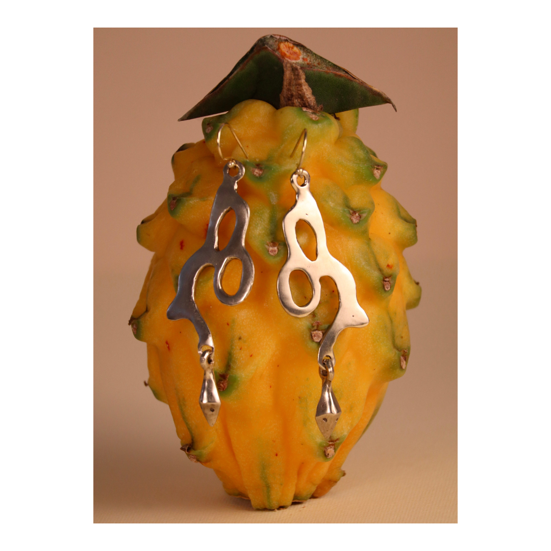A pair of Serpent Earrings by Madeleine Sculptural Jewellery, featuring an abstract silver dangle design, hang on a yellow dragon fruit with green-tipped scales against a neutral background.