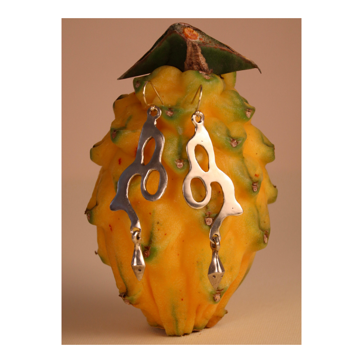 A pair of Serpent Earrings by Madeleine Sculptural Jewellery, featuring an abstract silver dangle design, hang on a yellow dragon fruit with green-tipped scales against a neutral background.