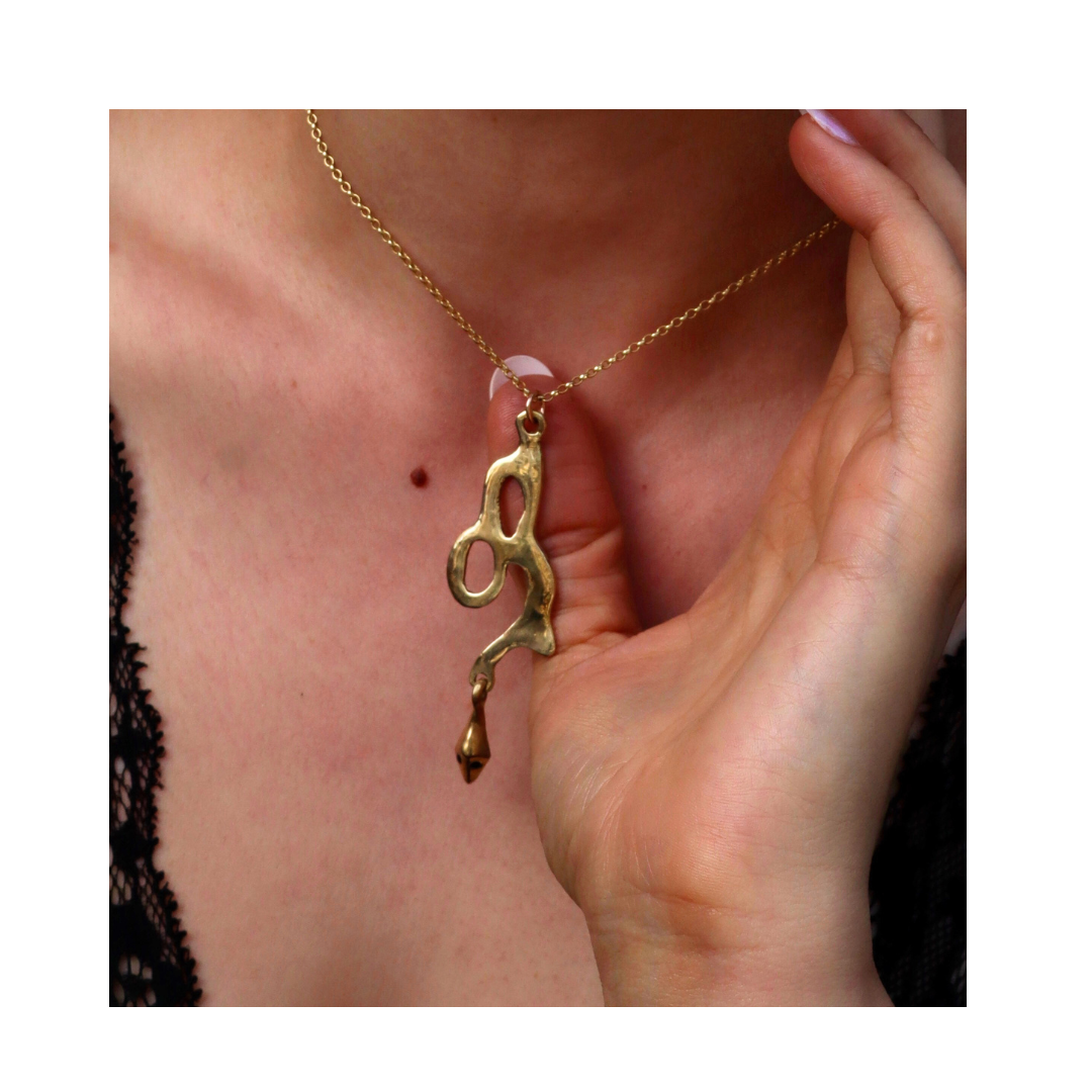 A hand holds the Serpent Pendant by Madeleine Sculptural Jewellery, an abstract-shaped necklace crafted from recycled brass, worn on a chain around the neck over a black lace-trimmed top.