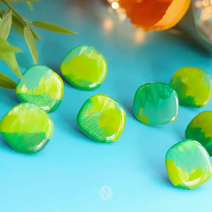 Close-up of small, rounded, translucent green and yellow glass beads scattered on a light blue surface. In the background, out of focus, are green leaves and an orange flower next to Naoi Shades of Green Stud Earrings with stainless steel earring posts.