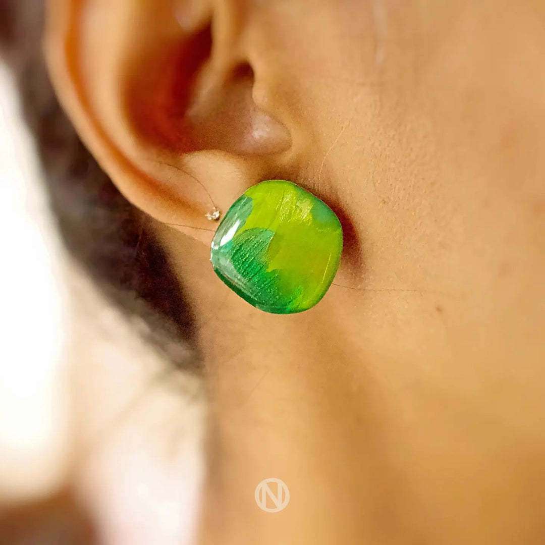 Close-up of a person's ear wearing Naoi's Shades of Green Stud Earrings with stainless steel earring posts. The brightly colored earring features a square-shaped, yellow and green design with a glossy finish. The person's hair is pulled back, highlighting the earring.