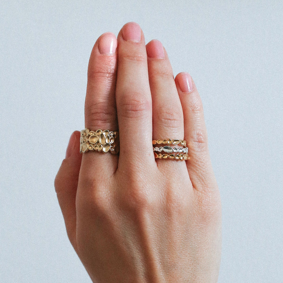 A hand with neatly manicured nails wearing multiple custom-made gold rings. One finger flaunts a thick, ornate ring, while another showcases two thinner rings, each with intricate designs. A Sara Gunn Shimmer Wide Ring Sterling Silver adds a touch of elegance against the plain, neutral background.