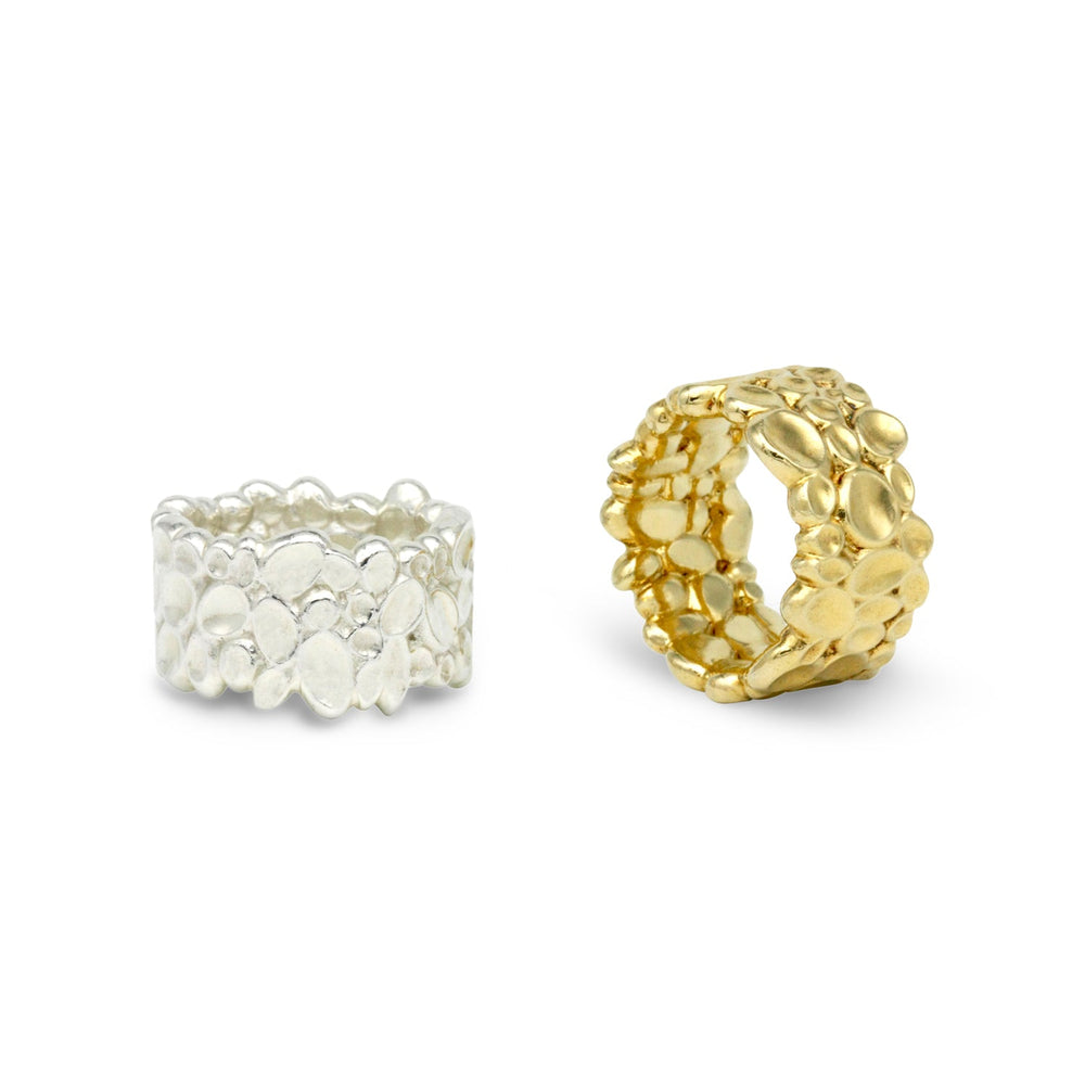 A pair of textured, custom-made rings is displayed against a plain white background. The ring on the left is crafted from a **Shimmer Wide Ring Sterling Silver by Sara Gunn**, while the ring on the right shimmers in gold. Both rings feature a pebble-like design with an irregular, organic texture.