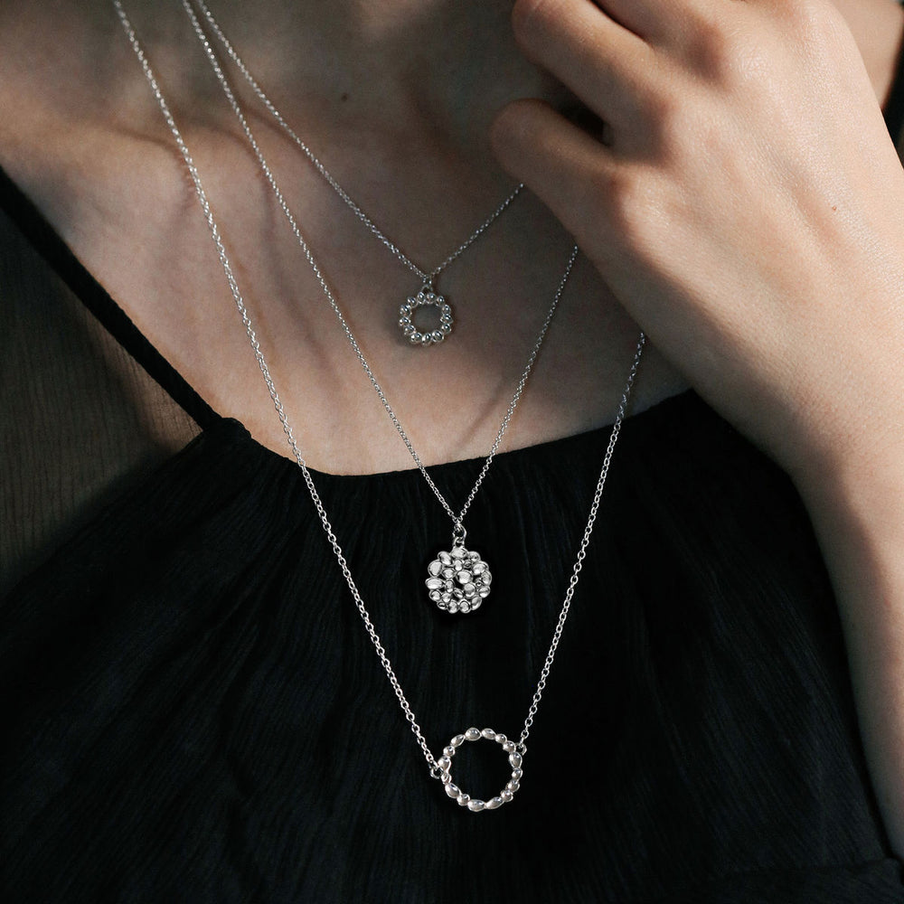 A close-up of a person wearing a black, sleeveless top. They are adorned with three layered sterling silver necklaces, each featuring a distinct circular pendant design. One hand gently touches the necklace chain, highlighting the delicate jewelry, including a Sara Gunn Shimmer Circle Pendant Sterling Silver.