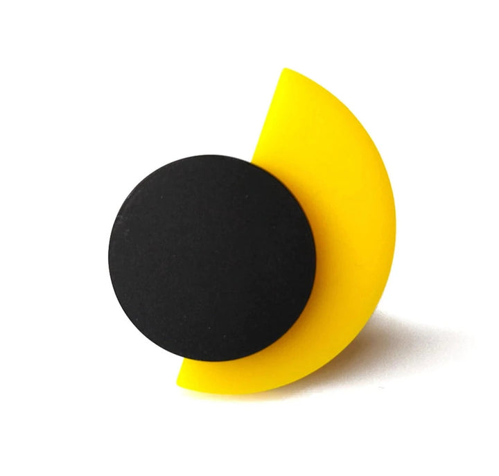 A minimalist, modern disk-like object featuring a black circle partially overlaid on a yellow semi-circle, creating a visually striking and balanced design. This handcrafted Suzan Ring from Alex + Svet, likely from a Paris studio, stands upright at an angle, casting a small shadow.