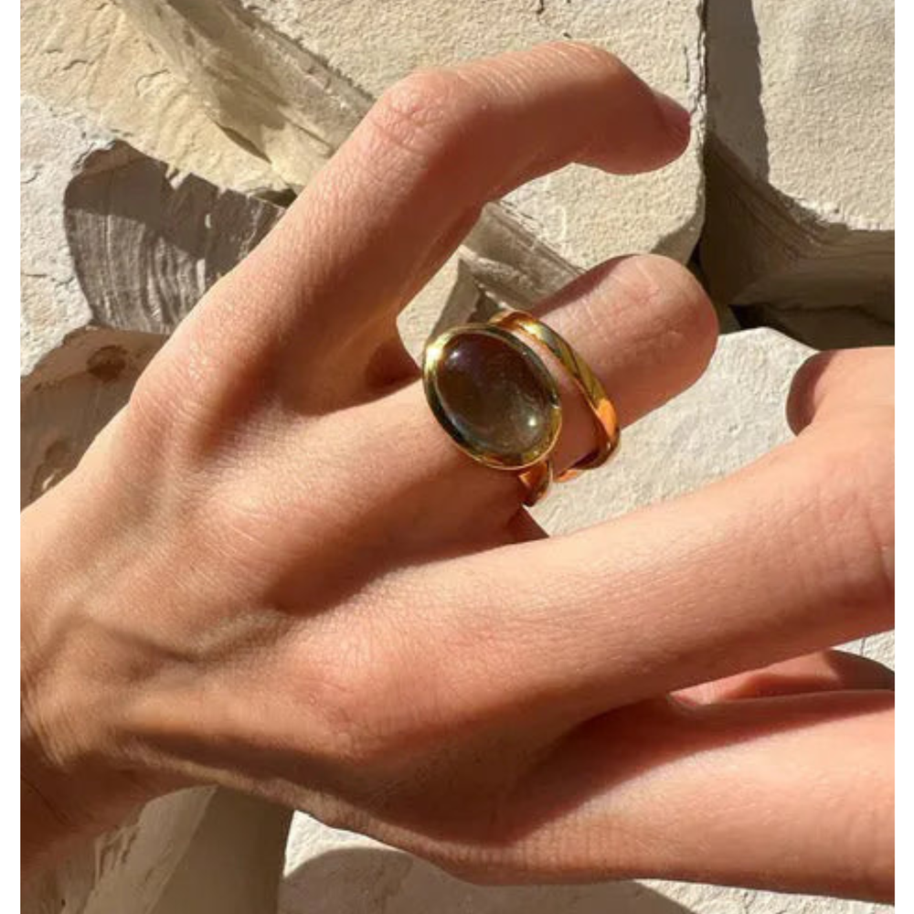 A hand with slender fingers wears the Shyla Sian Gold Plated Ring, featuring a large oval shape and reflective stone, shown against a textured, sunlit white stone background.