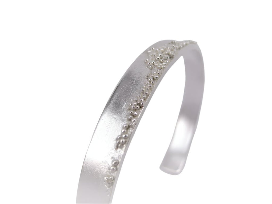 A sleek MarCucurella Silver Etrusc Bracelet with a smooth surface, adorned with small decorative beads along the top edge, creating an elegant and minimalist design reminiscent of Etruscan granulation against a plain background.