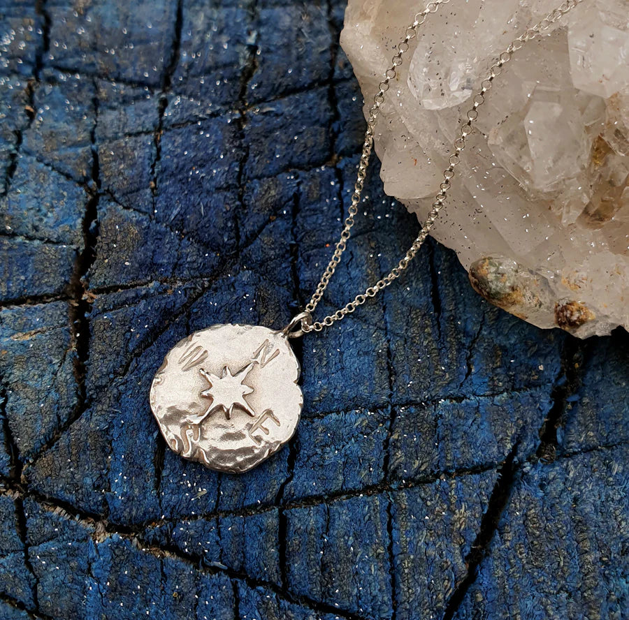 The Pale Raven Compass Coin Silver Necklace, handmade in Dublin, features a sterling silver pendant and is elegantly displayed beside a rock.