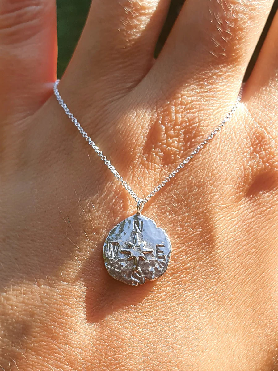 A close-up shows a hand wearing Pale Raven's Compass Coin Silver Necklace—handmade in Dublin with a round sterling silver pendant featuring N, S, E, and W. Sunlight highlights the fine compass details on both the necklace and hand.
