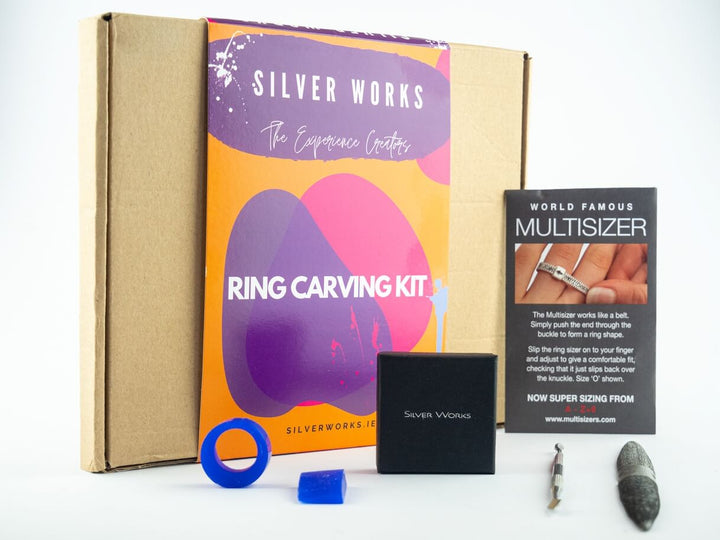 Image of a Silver Works - Ring Carving Kit by Silver Works including a cardboard box, instruction booklet, blue wax ring model, small black box, blue carving tool, and a Multisizer ring sizing strip card. The booklet has a purple and orange cover with the text "The Experience Creators." Perfect for jewellery artisans seeking a hands-on or virtual experience.