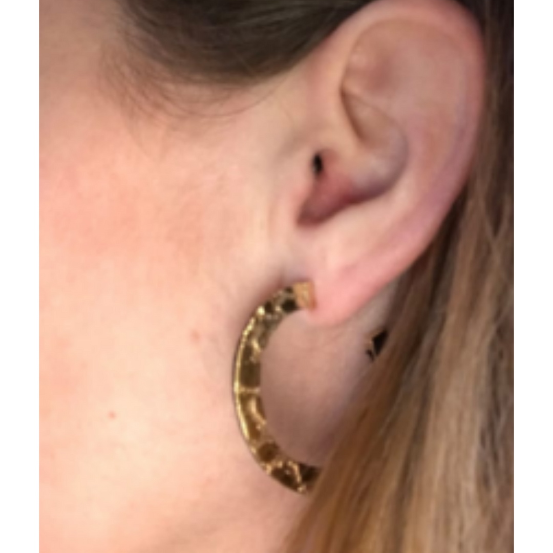 Close-up of a person's ear wearing Sara Gunn's Skin Etched Hoop Earring in gold-plated brass. The textured hoop stands out against partially visible light brown hair, with a softly blurred background.