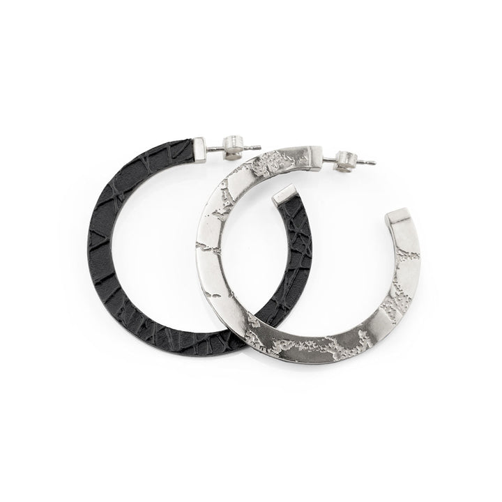 A pair of Sara Gunn Skin Etched Hoop Earrings; one earring is black with an embossed leather pattern, and the other is silver with a rough, uneven surface. Both earrings have a stud closure. The design features a mix of modern and rugged elements.