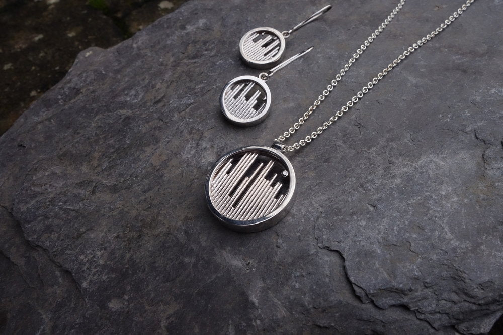 A set of matching Miriam Wade Skyline Drop Earrings Silver and a pendant necklace, displayed on a textured gray stone surface. Each piece showcases geometric circular drop earrings adorned with vertical lines and a small ball accent within the circle, featuring an Irish Hallmark guaranteed.