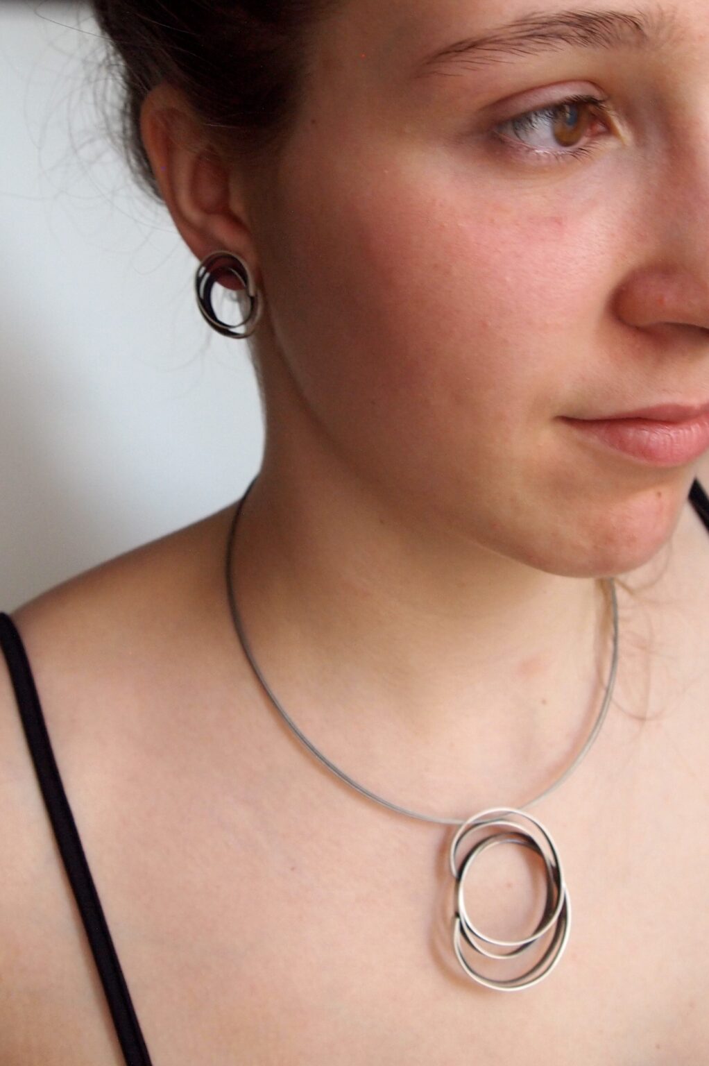 A person wearing a black spaghetti strap top is shown from the neck up. They have MarCucurella Small Mo Silver Earrings and a matching oxidized silver necklace with two interlocking rings. The background is plain and out of focus. Their hair is pulled back, and they are looking off to the side.