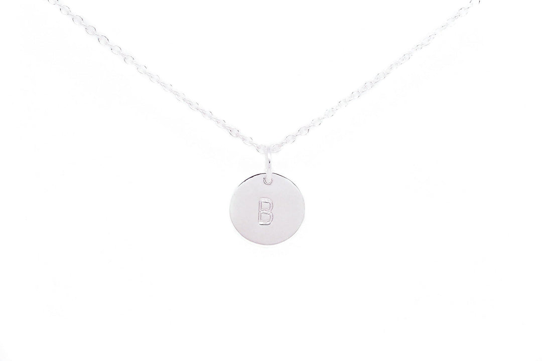 A delicate silver necklace featuring a circular pendant with the letter "B" hand stamped in the center. The Personalised Charms Personalised Small Disc Charm in Silver itself is an oval belcher chain, emphasizing the simplicity and elegance of the piece. The background is white, highlighting the jewelry.