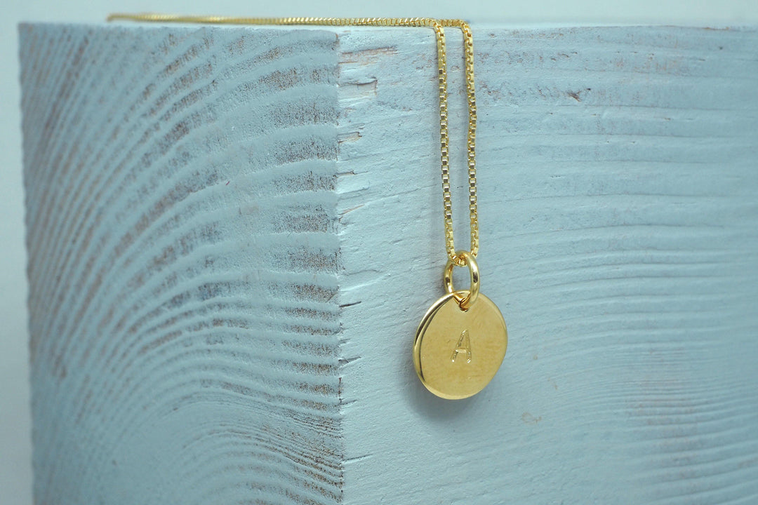 Personalised Charms Personalised Small Disc Charm in Gold with a hand-stamped charm engraved with the letter "A," draped over the edge of a textured piece of wood with a light grayish-blue hue.