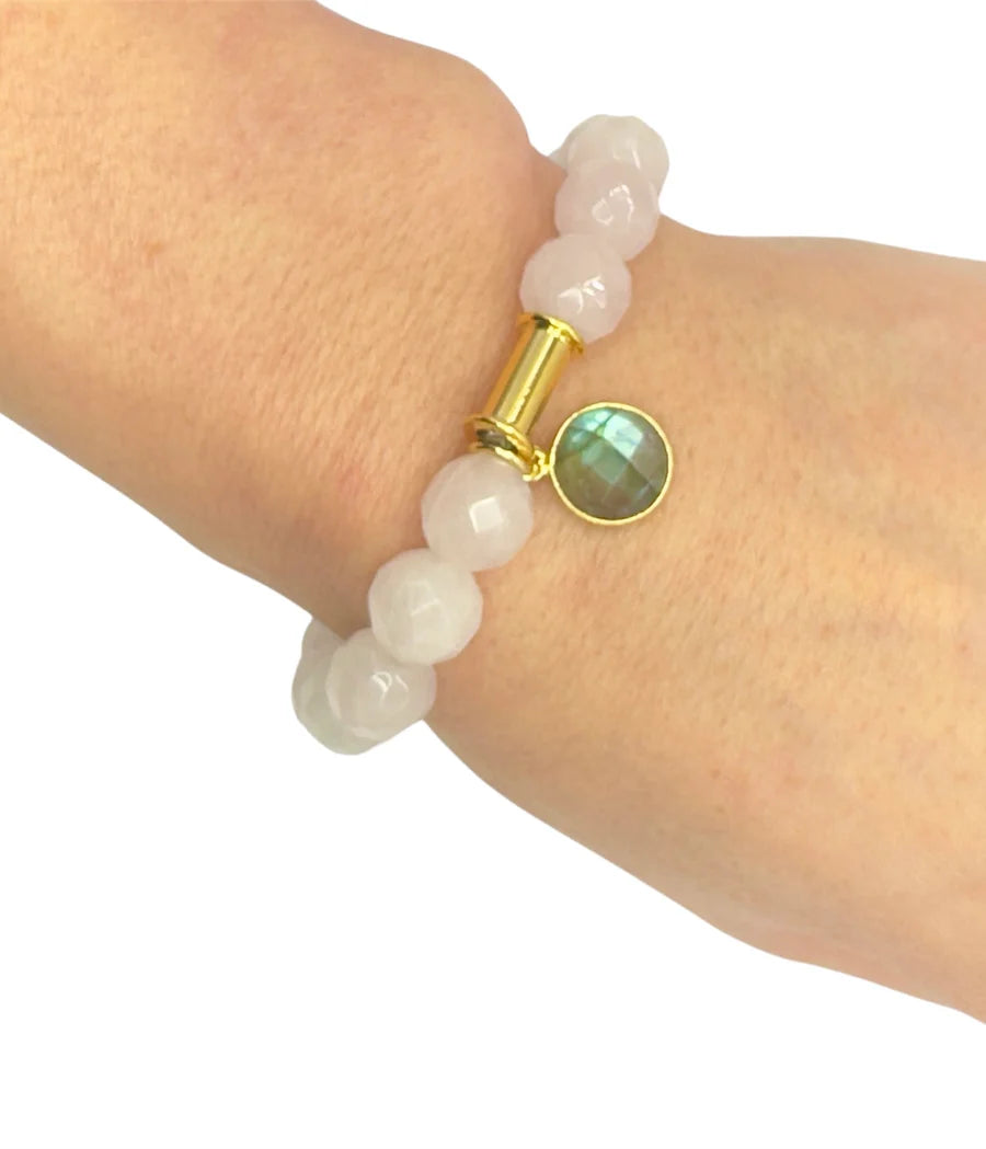 A hand wears the Aria V Soulstone Rose Quartz and Labradorite Bracelet—round, faceted pale pink beads, a gold cylinder accent, and a green labradorite charm in gold—an elegant piece of spiritual jewelry.