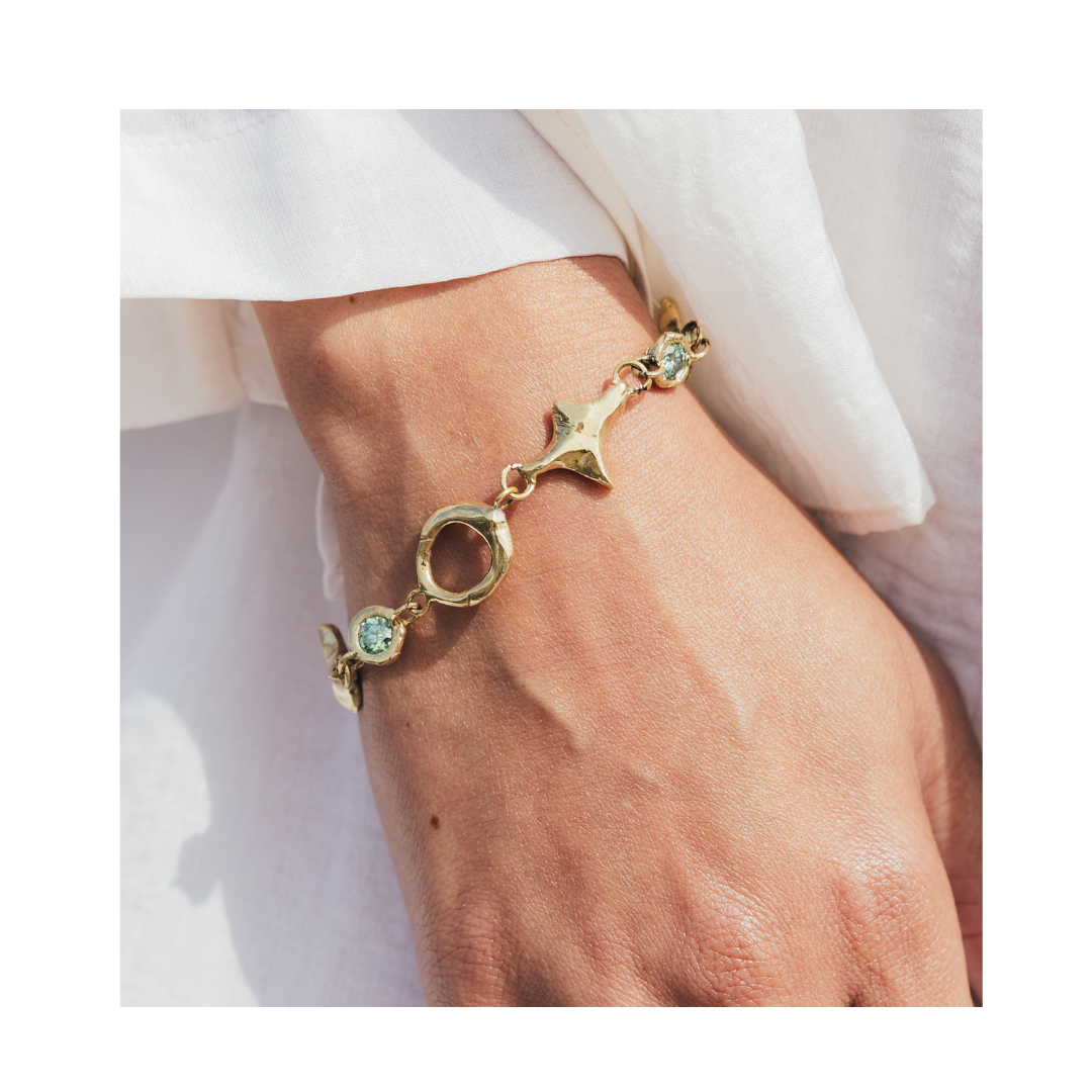 A close-up of a hand wearing the Madeleine Blaine Spark Bracelet—a gold bracelet featuring circular and star-shaped links adorned with light blue stones—highlights its vibrant connections and energy against a backdrop of white clothing.