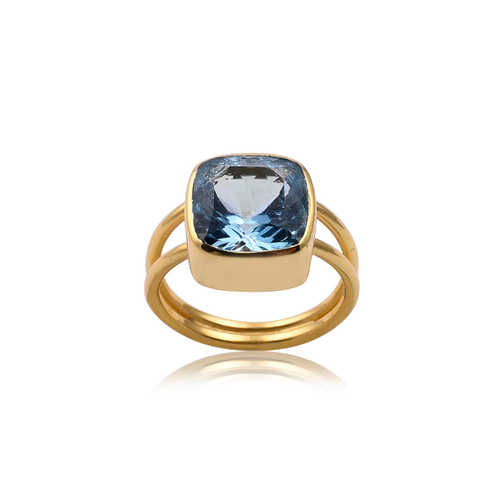 A Square Blue Quartz Gold Ring with a wide band and open-style shank features a large, cushion-cut Blue Quartz gemstone in the center. The gemstone is set in a simple bezel setting, accentuating its vibrant hue. This piece of handmade jewellery by Watermelon Tropical is positioned upright against a white background.