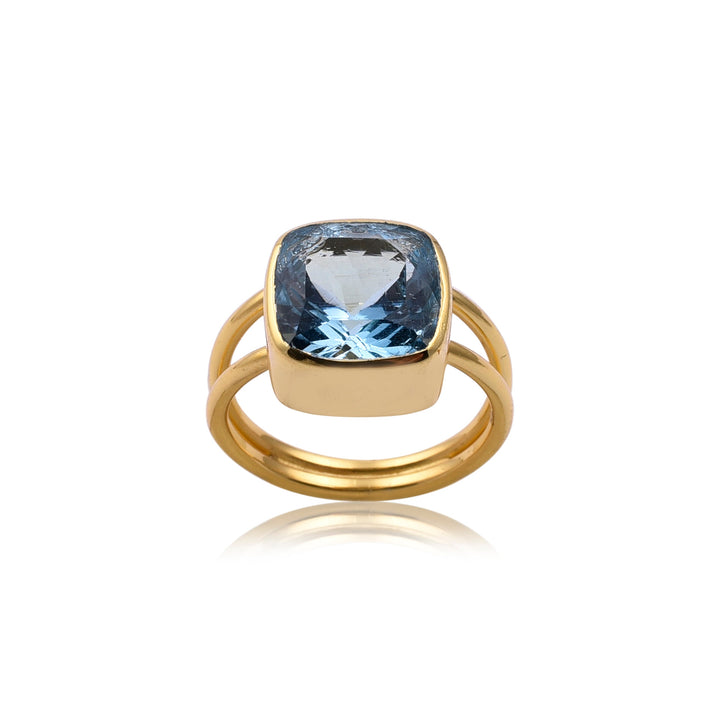 A Square Blue Quartz Gold Ring with a wide band and open-style shank features a large, cushion-cut Blue Quartz gemstone in the center. The gemstone is set in a simple bezel setting, accentuating its vibrant hue. This piece of handmade jewellery by Watermelon Tropical is positioned upright against a white background.