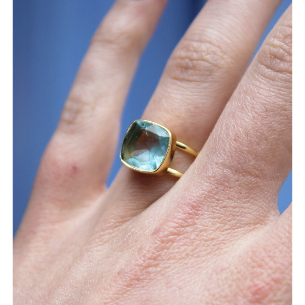 A close-up of a hand wearing Watermelon tropical’s Square Blue Quartz Gold Ring, featuring a large, square-cut blue quartz gemstone. The background is blurred and blue-toned.