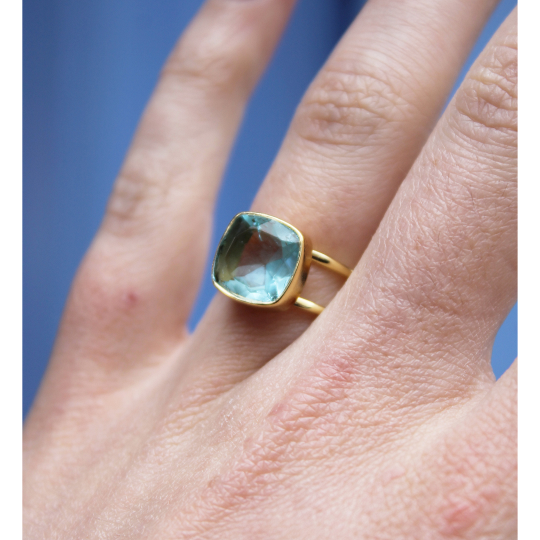 A close-up of a hand wearing Watermelon tropical’s Square Blue Quartz Gold Ring, featuring a large, square-cut blue quartz gemstone. The background is blurred and blue-toned.
