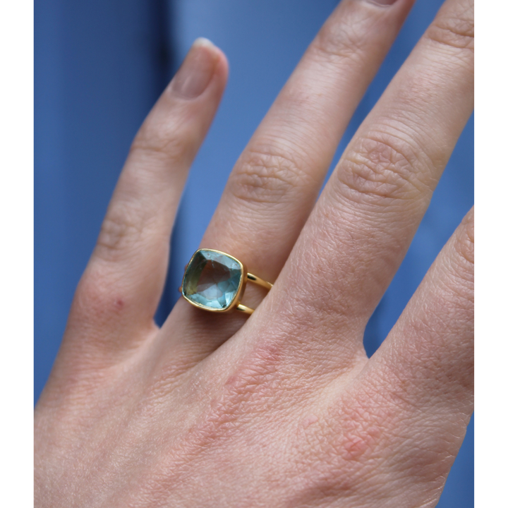 A hand displays the Watermelon tropical Square Blue Quartz Gold Ring, featuring a large, square light blue quartz set in gold vermeil, shown against a soft blue background.