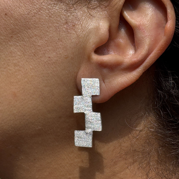 A close-up of a person’s ear wearing Soufana Attia’s Stairway to Heaven Earrings - Large showcases three stacked, glittery squares in a geometric silver design. The blurred background accentuates the earrings’ bold texture and shine.