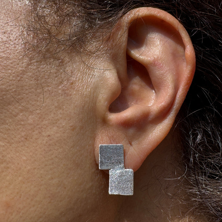 Close-up of a person's ear wearing Soufana Attia's Stairway to Heaven Earrings - Small: asymmetric geometric silver earrings with two overlapping matte-finish squares. Person's skin and hair are visible in the background.