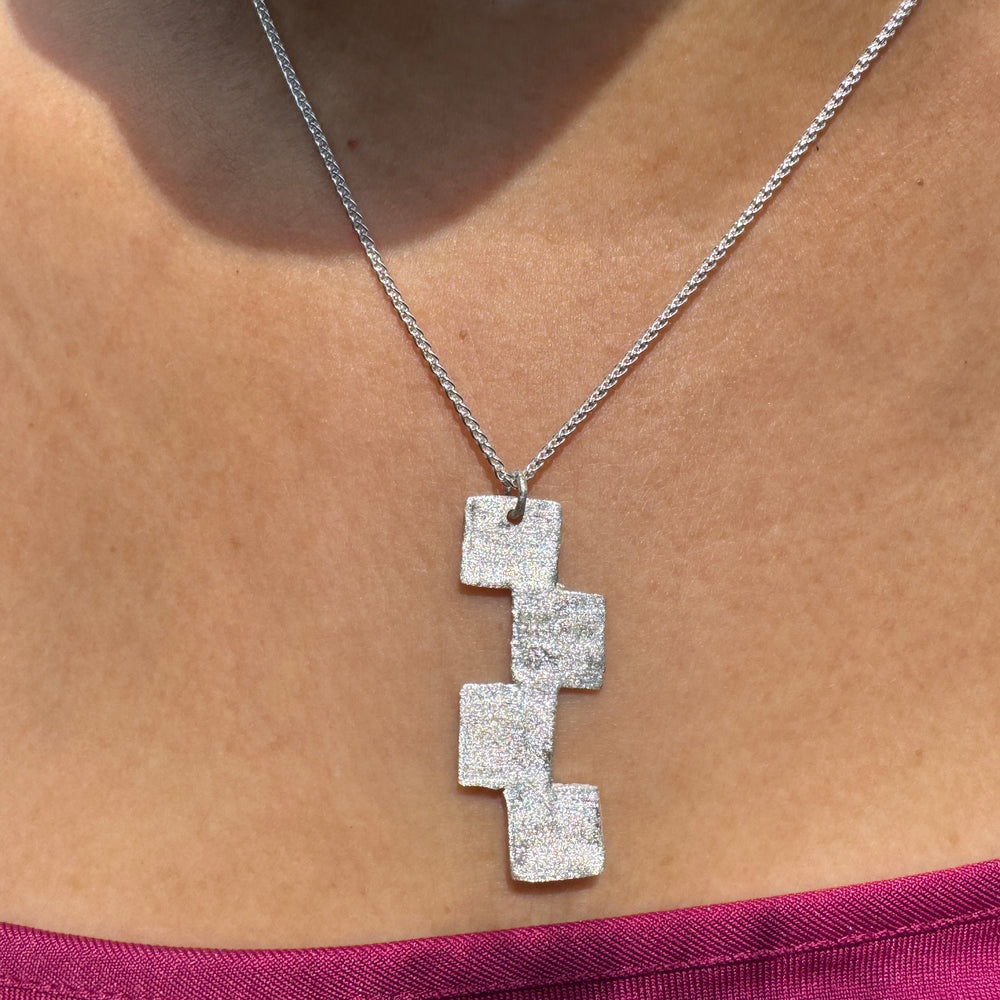 A close-up of a person in a magenta off-the-shoulder top wearing the Soufana Attia "Stairway to Heaven Pendant"—a silver necklace featuring three stacked, glittery squares that evoke the idea of an upward journey.