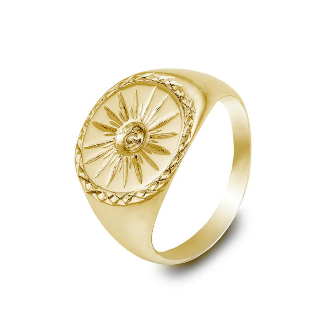The Cosmic Boulevard Starburst 9ct Gold Signet Ring features an embossed sunburst design with a central engraved face. The band is smooth and polished, and the round face of this handmade signet ring has additional intricate detailing around the edge.