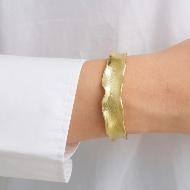 A person in a white long-sleeve shirt showcases the Watermelon tropical Statement Bangle—gold plated brass with wavy edges—on their wrist against a plain background.